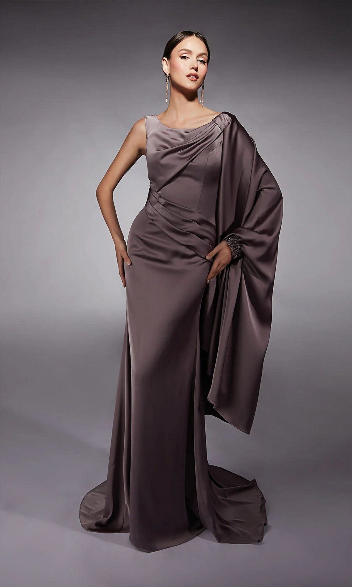 A woman poses in the Alyce 27722 Long Formal Dress—a floor-length, taupe one-shoulder satin gown with draped detailing and a fitted bodice—against a plain gray background.